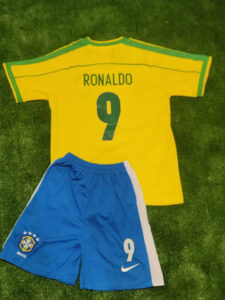 s-l960_11 | UAE SPORT STORE Yellow Brazil soccer jersey with 'RONALDO' and number 9 laid over blue shorts on grass, celebrating a football kit set up.