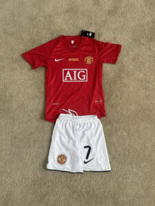 s-l960_2 | UAE SPORT STORE Red Manchester United home jersey with AIG sponsor and club crest, laid over white shorts on a beige carpet.