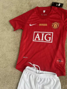 s-l960_3 | UAE SPORT STORE Red Manchester United jersey on beige carpet, with AIG sponsor, white Nike swoosh, and club crest beside the tag, paired with white shorts.