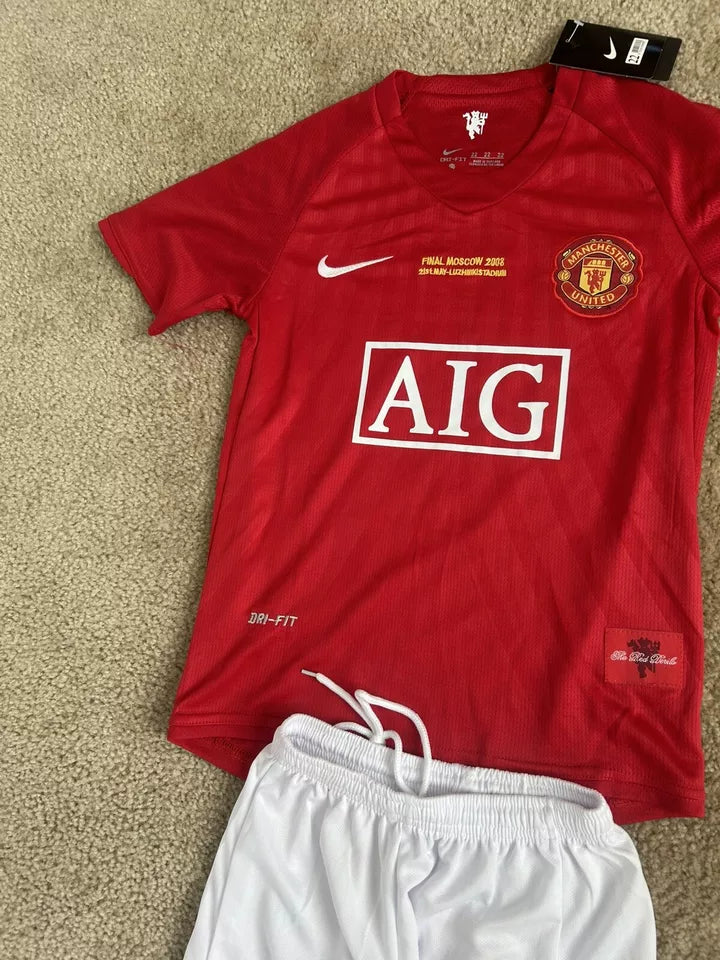 s-l960_3 | UAE SPORT STORE Red Manchester United jersey on beige carpet, with AIG sponsor, white Nike swoosh, and club crest beside the tag, paired with white shorts.