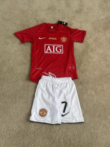 s-l960_5 | UAE SPORT STORE Manchester United red home jersey with AIG sponsor and crest, laid on a carpet beside white shorts with a large '7' and Nike swoosh.