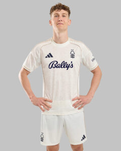 Male model in a white soccer jersey and shorts, standing with hands on hips against a gray background; Bally's sponsor logo on chest and a crest on the left.