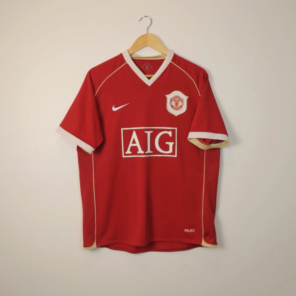 Red Manchester United football jersey on a wooden hanger against a pale wall, displaying the team crest and AIG sponsor logo.