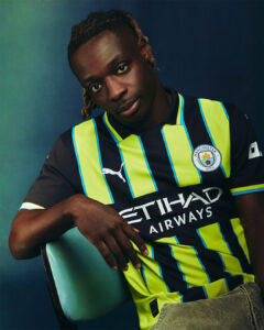 Young man seated, wearing Manchester City football jersey with neon yellow-green stripes, looking at the camera.