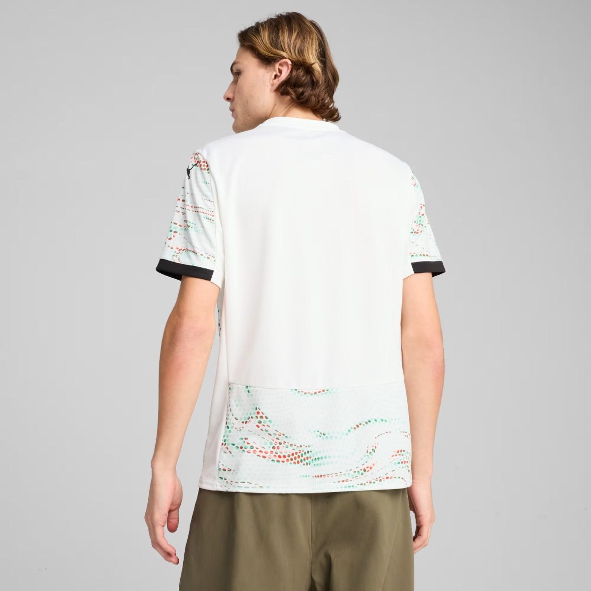 Back view of a person wearing a white short-sleeve shirt with colorful patterned sleeves and olive shorts, standing against a neutral gray background.