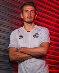 Portrait of a man in a white Sheffield United jersey with arms crossed against a red-lit striped wall.