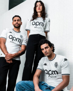 Three models wearing white APM Monaco football jerseys pose against a white tiled wall, looking confident.
