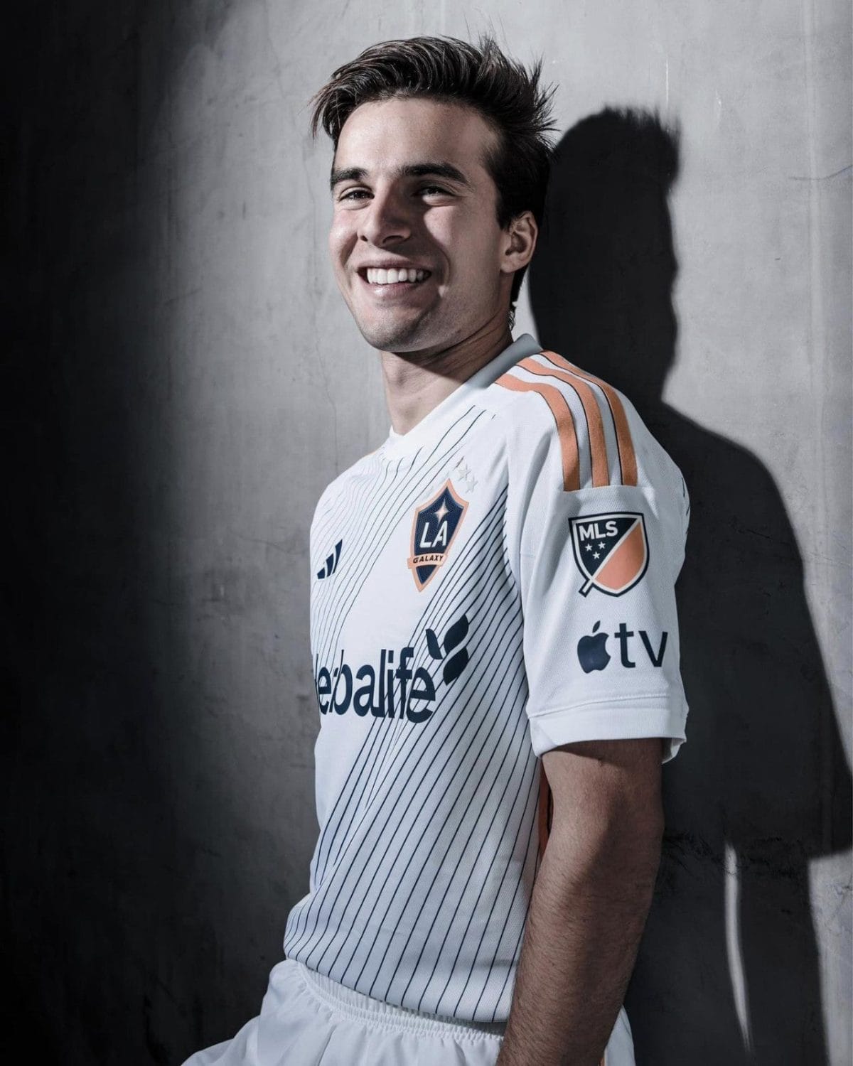 Smiling male soccer player in an LA Galaxy jersey leaning against a concrete wall.