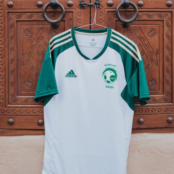 White and green Adidas Saudi football jersey hanging on a hanger in front of a carved wooden door; features SAFF crest and Adidas logo.
