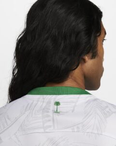 Back view of a person with long black hair wearing a white jersey with a green collar and a small palm tree logo at the back neck area, viewed from behind, on a light background.