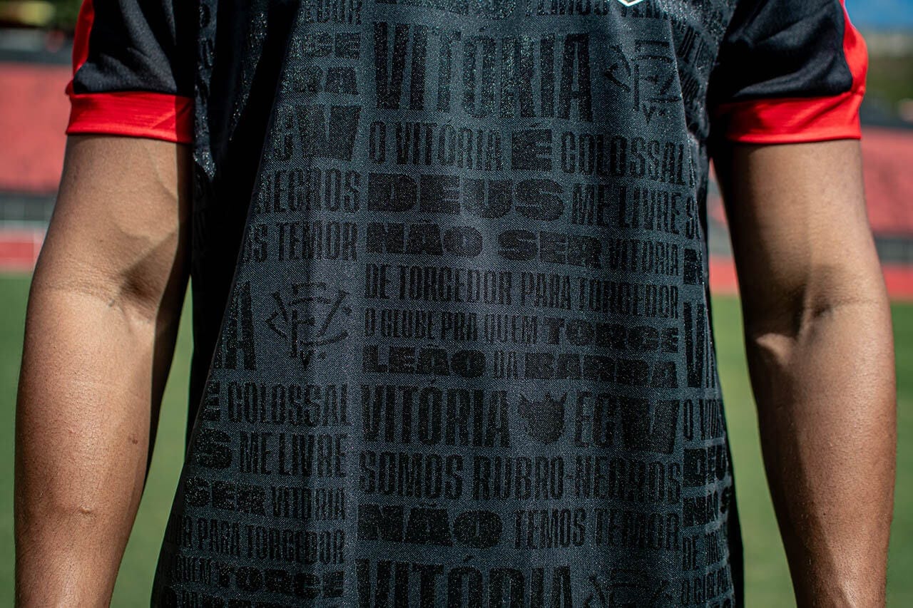 sdhhd | UAE SPORT STORE Person wearing a black athletic jersey with red sleeve cuffs on a football field; shirt has bold black text patterns and symbols.