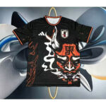 Black Japan soccer jersey with a bold red demon mask graphic, Adidas logo, and orange trim, hanging on a hanger against a colorful abstract background.