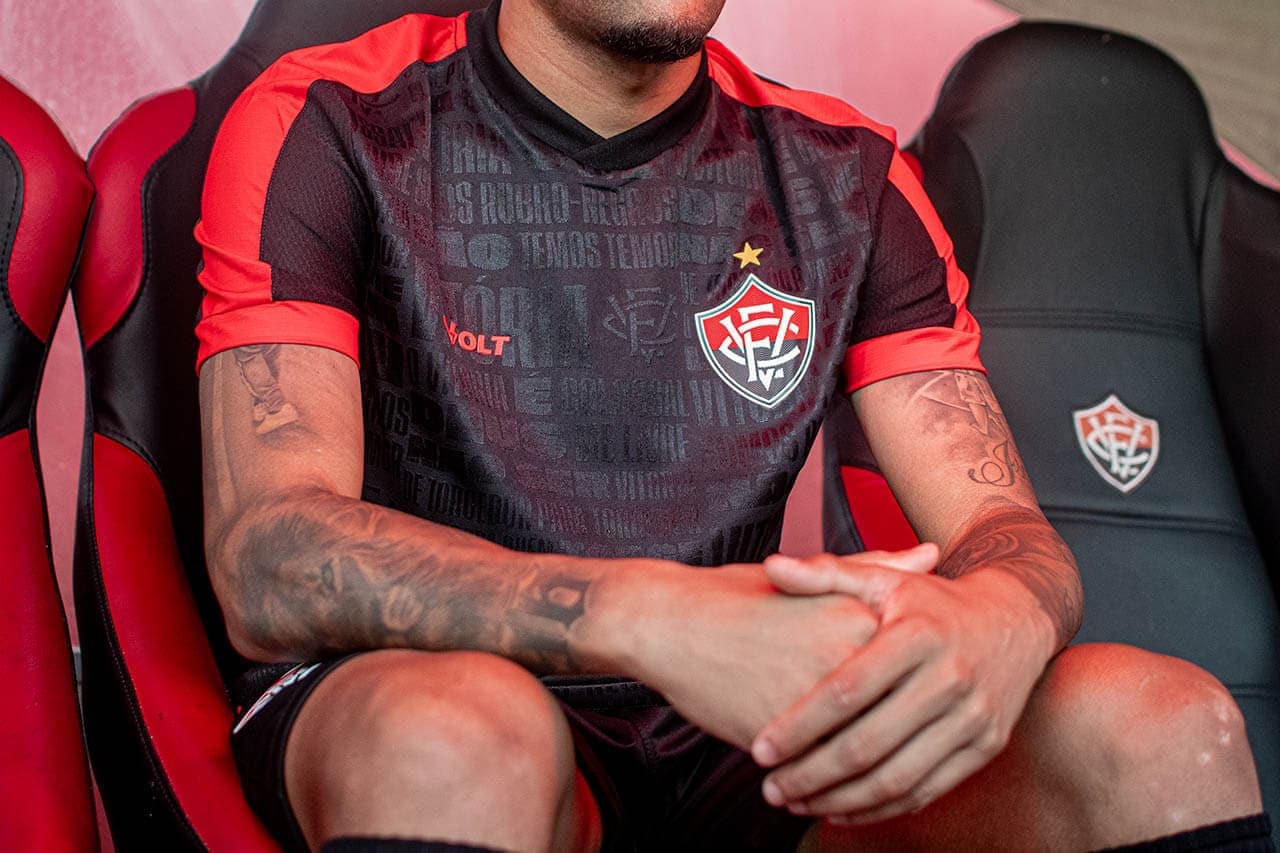 sgsgf | UAE SPORT STORE Athlete in a black and red jersey sitting on a bench with folded hands, displaying a crest with a star on the chest and visible tattoos on arms.