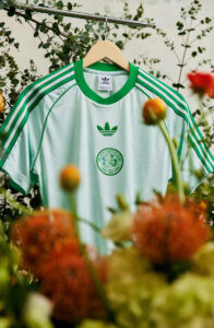 shirt_homepage_banner_mobile_750x1150_adf4492d-b009-4571-bd7d-e74b3e03f6cf | UAE SPORT STORE Light green Celtic FC jersey on a wooden hanger, hung outdoors among blurred flowers and greenery.