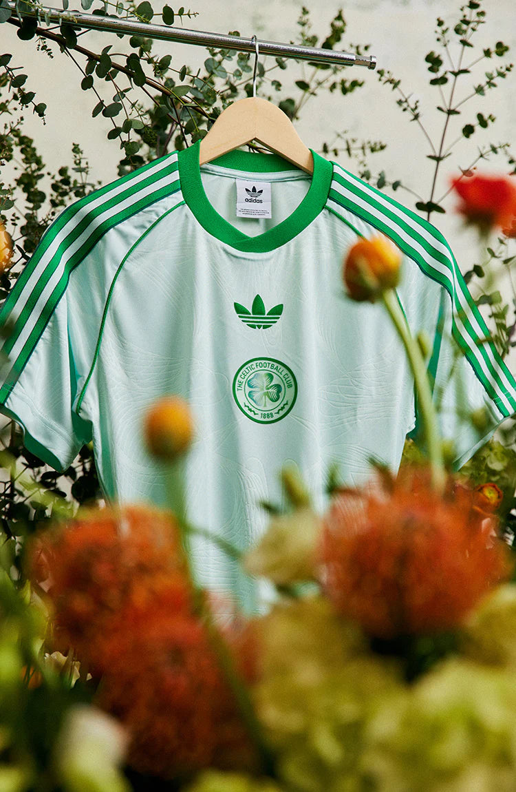 shirt_homepage_banner_mobile_750x1150_adf4492d-b009-4571-bd7d-e74b3e03f6cf | UAE SPORT STORE Light green Celtic FC jersey on a wooden hanger, hung outdoors among blurred flowers and greenery.