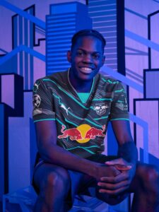 Portrait of a smiling man in a dark teal jersey with the Red Bull logo, seated against a blue geometric mural backdrop.