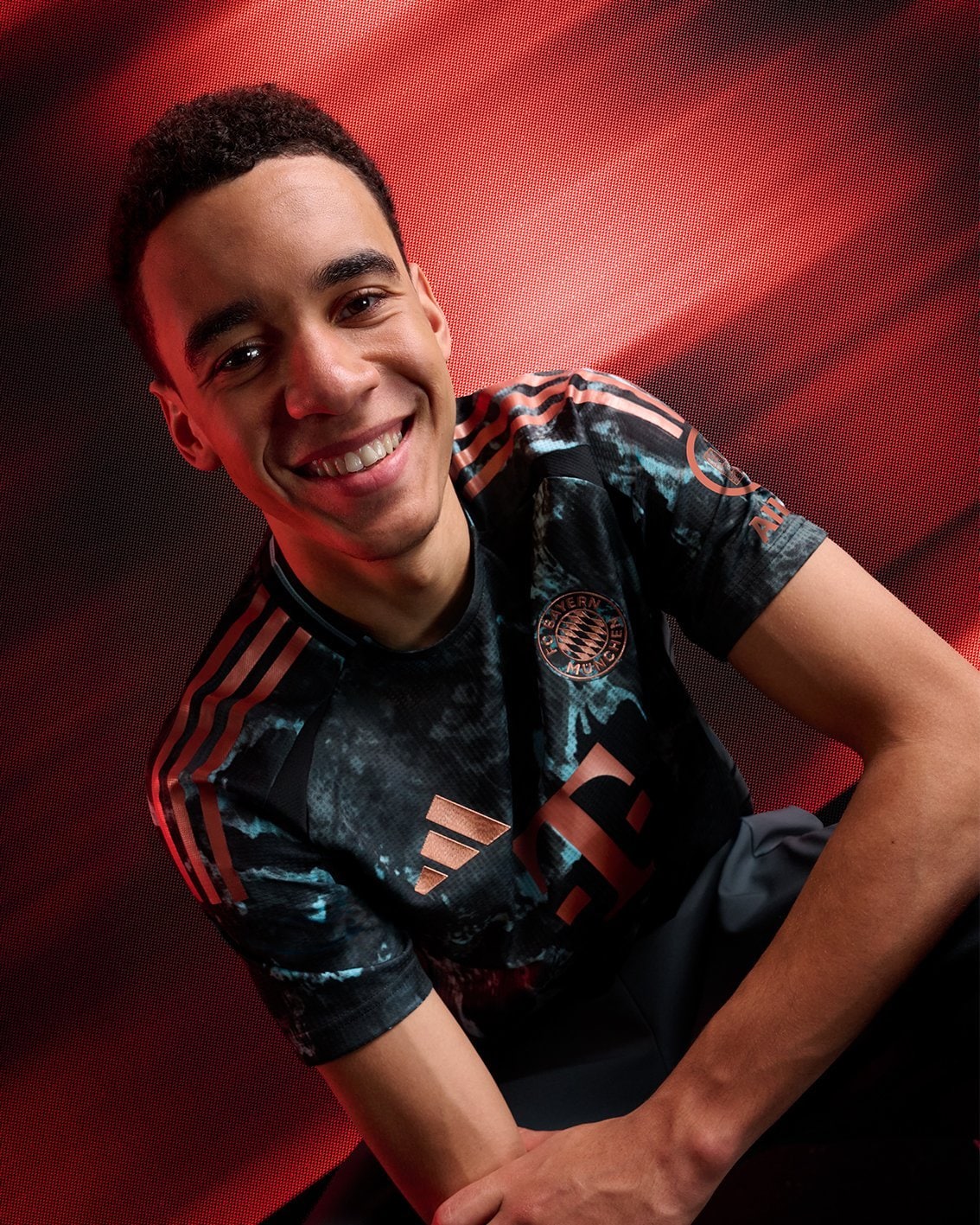 sjDbNmiDgWNGf0U | UAE SPORT STORE Smiling man in a Bayern Munich jersey posing against a red textured backdrop.