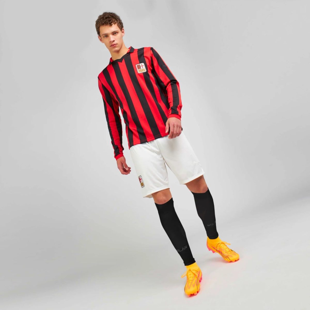 Soccer player in a red-and-black striped jersey, white shorts, black socks, and bright orange cleats, posing on a gray backdrop.