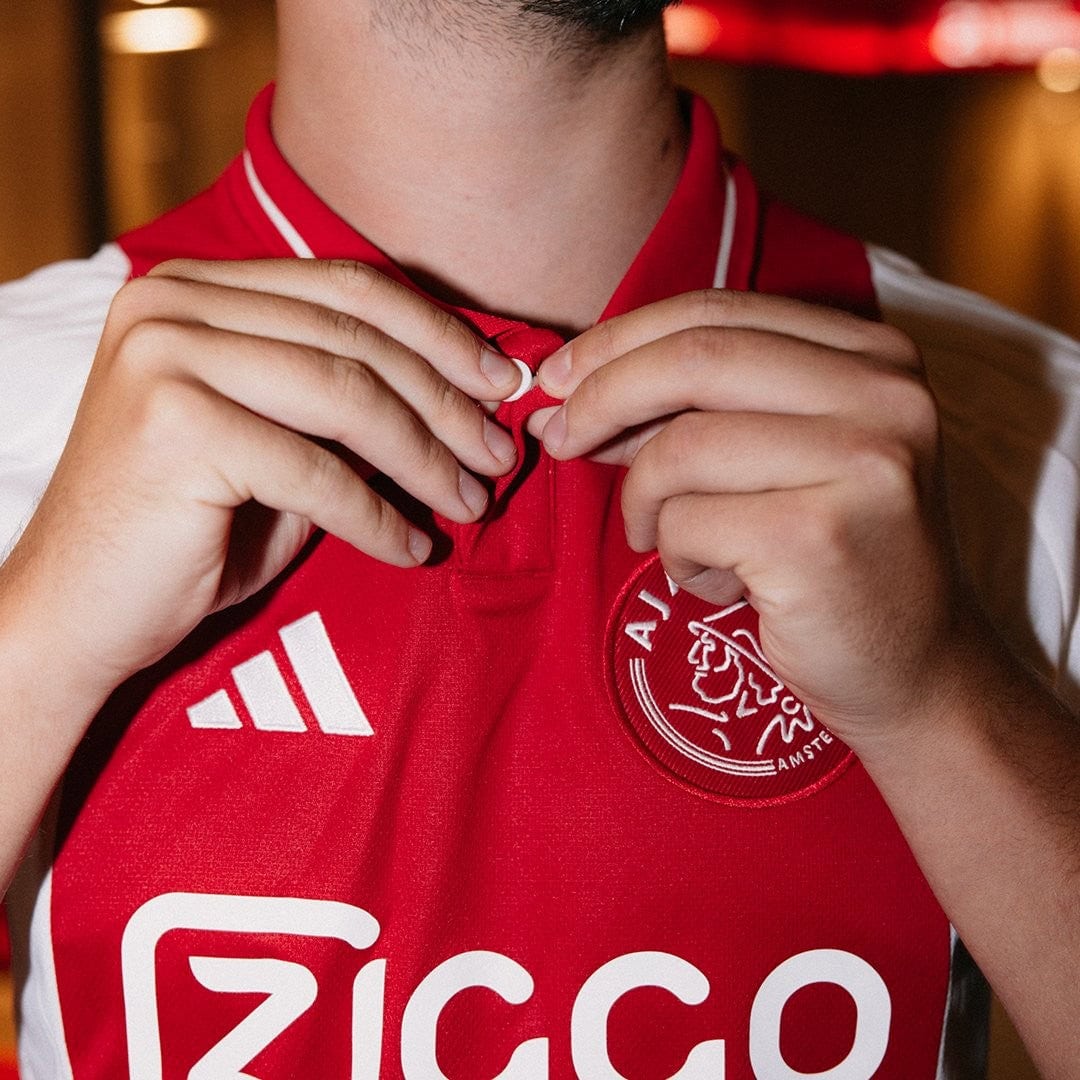 svJxrmyw2YO0D9N | UAE SPORT STORE Close-up of a person adjusting the red Ajax football jersey collar, with the Ziggo sponsor and club crest visible on the chest.