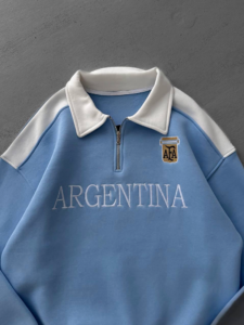 Light blue sweatshirt with white collar and a half-zip, featuring an 'ARGENTINA' wordmark across the chest and a small crest.