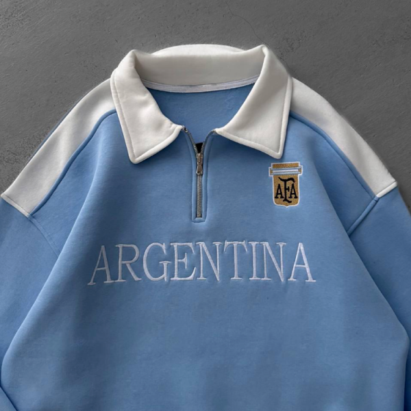 Light blue sweatshirt with white collar and a half-zip, featuring an 'ARGENTINA' wordmark across the chest and a small crest.