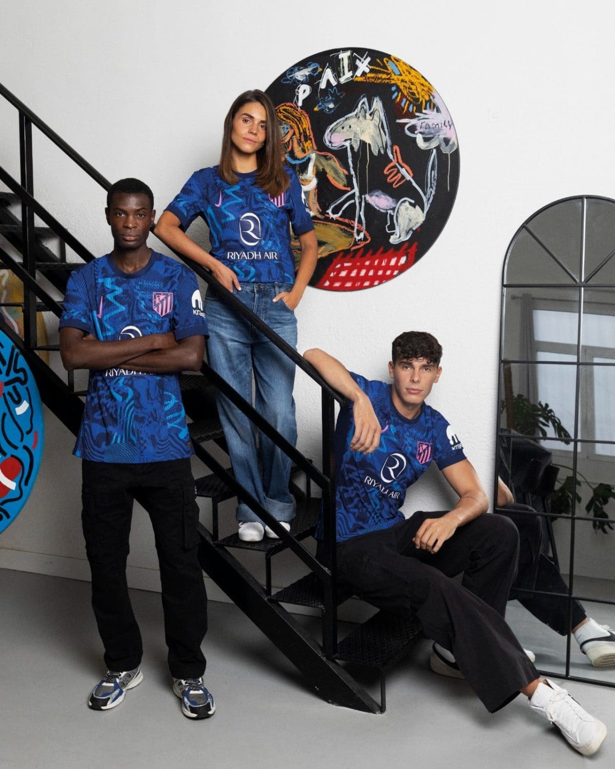 Three young adults in blue Riyadh Air jerseys posing on a black staircase beside a colorful circular mural behind them.