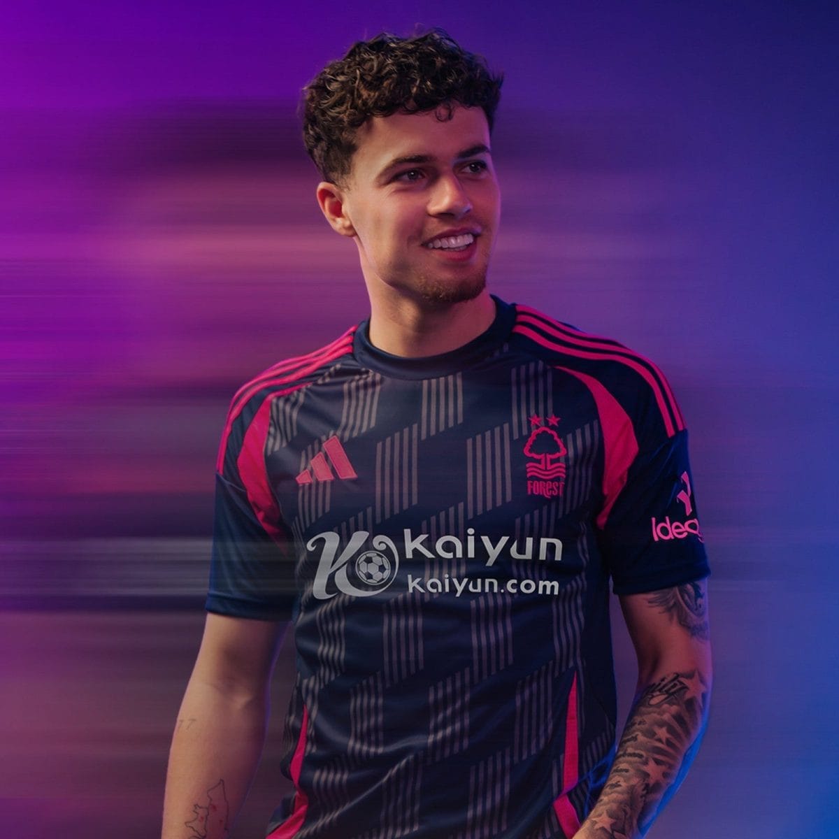 Young man in a dark soccer jersey with pink accents smiling, looking to the left, with a purple-pink motion-blur background.