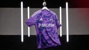 tIXdkIZRUfQmXtW | UAE SPORT STORE Fulham FC purple camouflage soccer jersey with SBOTOP sponsor hanging on a hanger in a studio setting.