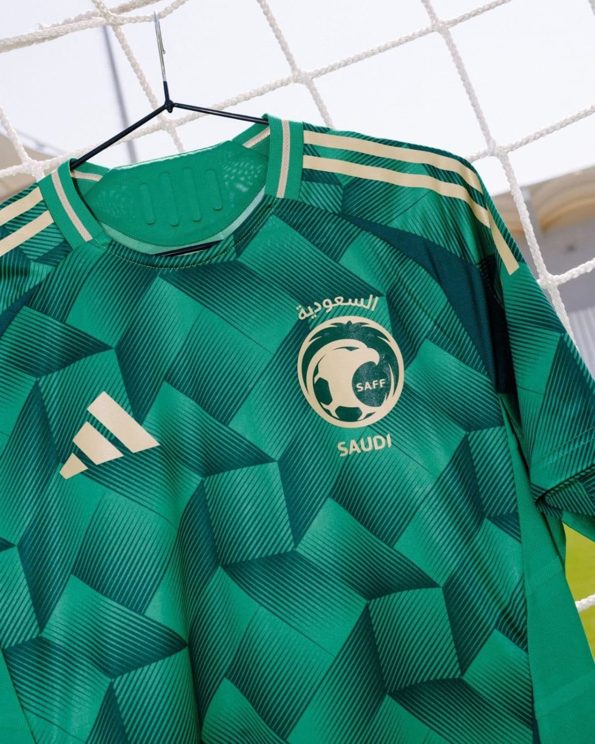 Green Saudi national football team jersey on a hanger, showing the SAFF emblem and Adidas three stripes on the shoulders.