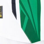 Close-up of a white mesh sports jersey with green panels and a small black-and-yellow brand patch on the left side, showing stitching detail.