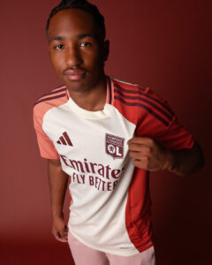 Young man wearing an OL Lyonnais soccer jersey, pulling the shirt to show the crest against a red backdrop.