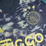 Close-up of an Ajax Amsterdam football jersey showing the circular club crest with three gold stars above it on a dark, speckled fabric.