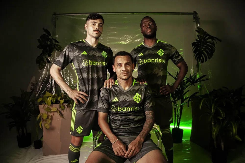 tj62CuvvHo9Eu0D_988bf35b-4130-4771-9447-eef0f09812ba | UAE SPORT STORE Three male soccer players pose for a team portrait in dark camouflage jerseys with neon green trim and 'banrisul' sponsor logos, in a plant-filled studio.