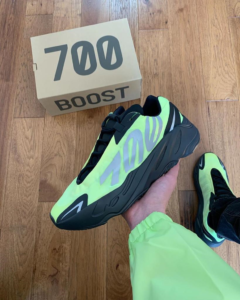 Neon yellow-green Yeezy Boost 700 sneaker held in hand over a wooden floor with a '700 BOOST' box in the background.