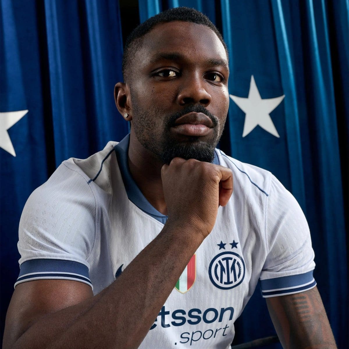Close-up portrait of a Black man in an Inter Milan jersey, resting his chin on his hand against a blue backdrop with stars.