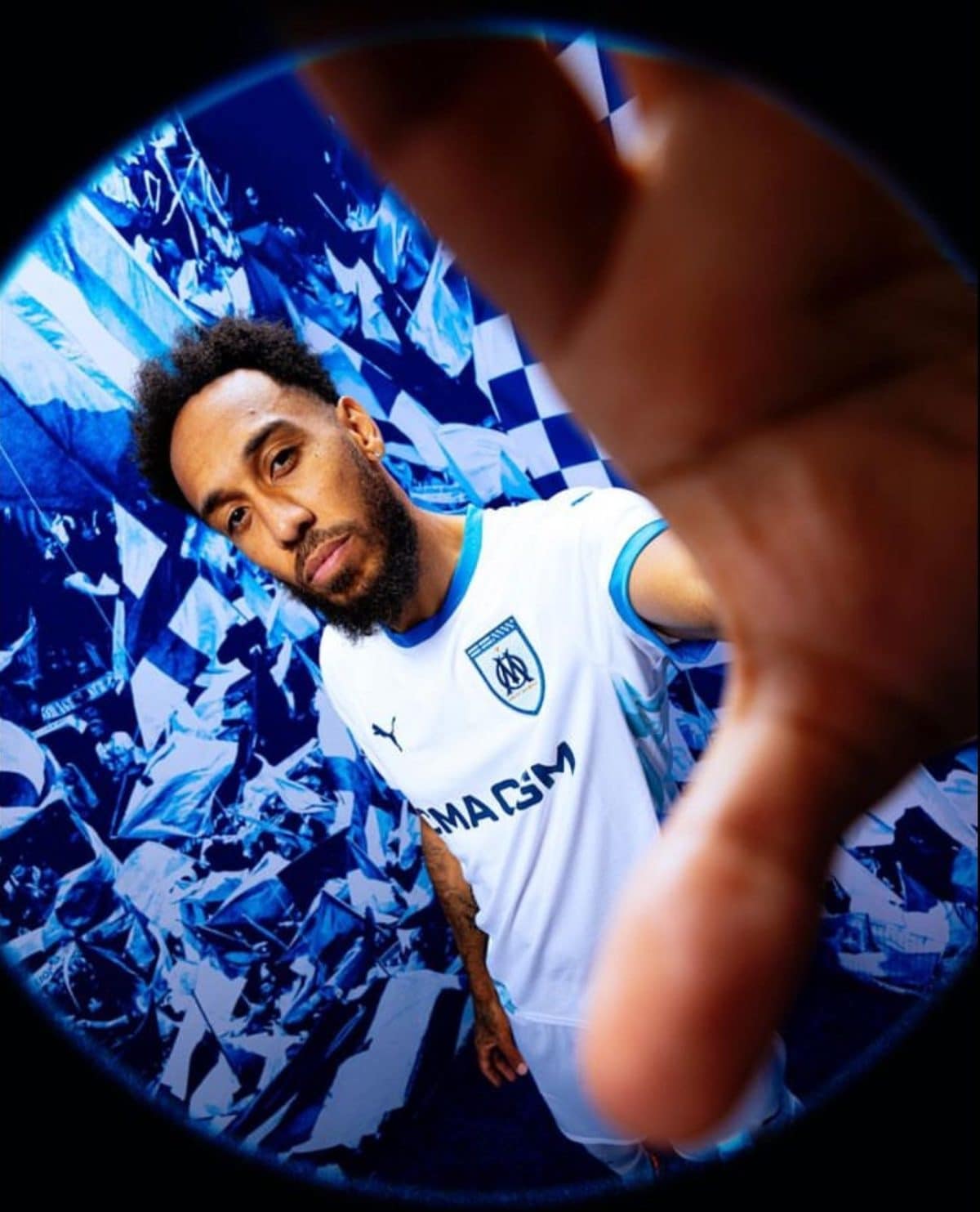 A man in a white and blue soccer jersey reaches toward the camera, with a blue geometric background and a blurred foreground hand nearby. The shot feels like a dynamic action moment.