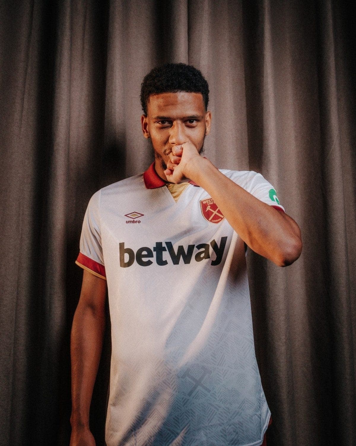 A football player wearing a West Ham jersey with 'betway' sponsor, standing against a dark curtain, with his hand near his mouth in a thoughtful pose.