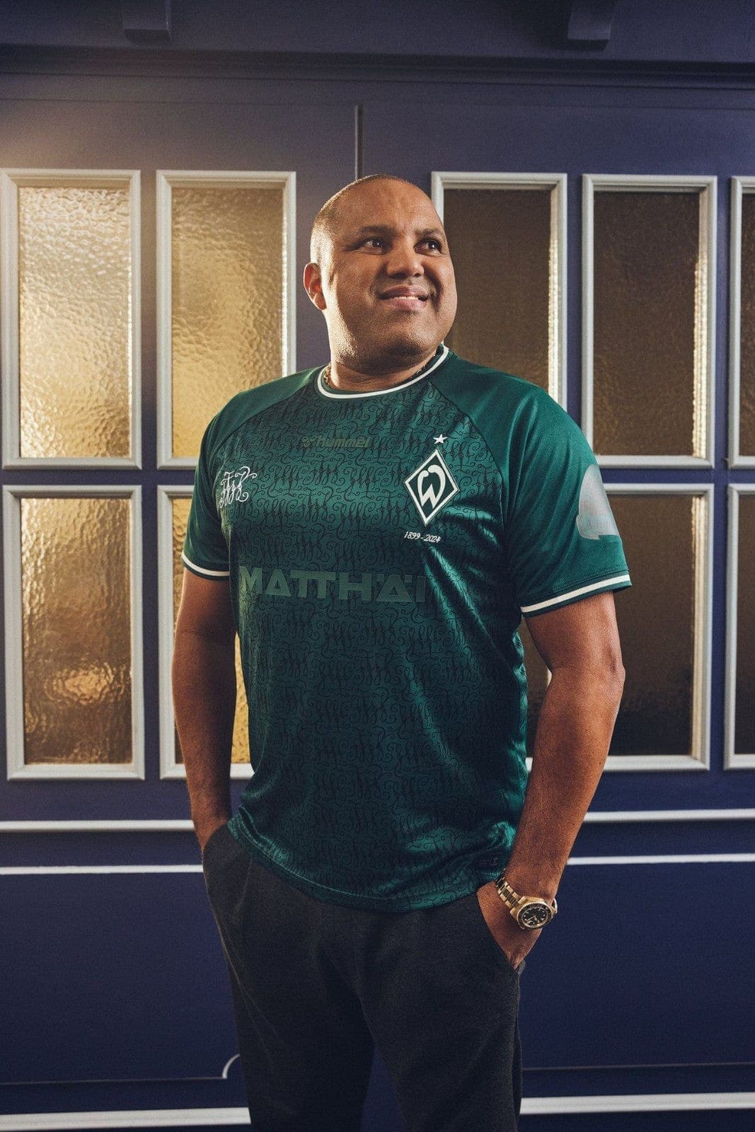 uBZX7IipoLEaUDD | UAE SPORT STORE Smiling man in a teal sports jersey with a crest stands against a blue wall with glass-panel windows behind him.