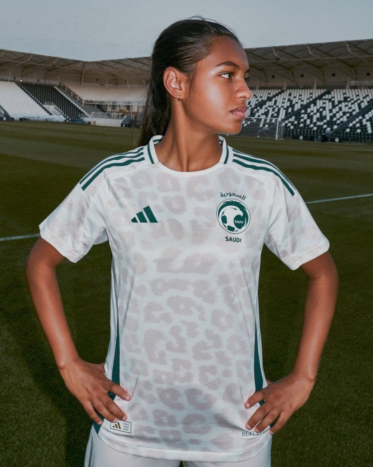 Young woman soccer player wearing a white Saudi jersey with SAFF emblem, standing on a football field with stadium seating in the background, hands on hips.