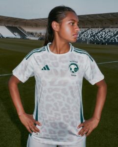 Young woman soccer player wearing a white Saudi jersey with SAFF emblem, standing on a football field with stadium seating in the background, hands on hips.