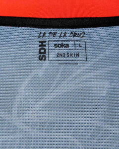 Clothing tag showing brand text 'SODA' vertically, with 'soka' logo, size L, and '2ND SKIN' on a striped fabric background.