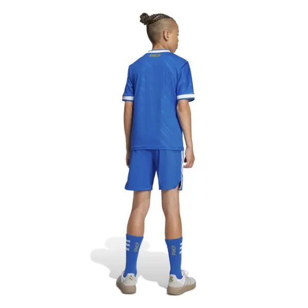 Back view of a child wearing a blue youth soccer uniform (jersey, shorts, and long blue socks) with beige cleats, standing on a white background.