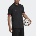 Male model wearing a black Adidas soccer jersey with red trim, holding a white soccer ball.