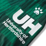 ujjkjkj | UAE SPORT STORE Close-up of a green jersey with large white 'UH' lettering and 'University Of Hertfordshire' text, plus a Kelme paw logo.