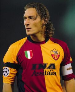 Soccer player in a red and yellow striped jersey with team crest, UEFA patch on the sleeve, looking to the left on the field