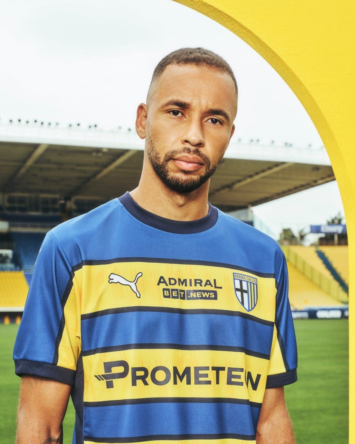 Portrait of a man in a blue and yellow soccer jersey standing on a stadium field under a yellow arch, looking at the camera, sponsorship logos visible on chest.