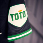 utHHP4GG2se4C1u | UAE SPORT STORE Close-up of a black sleeve with a white shield patch reading TOTO in green letters and a small orange accent above, bordered in green.