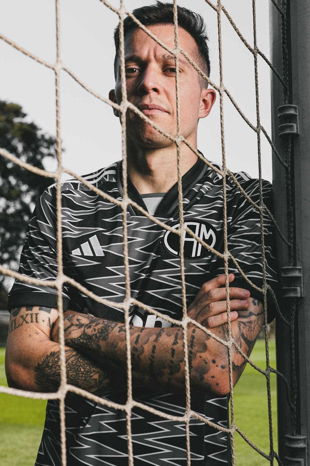 uuy1FI4DGooEWfG | UAE SPORT STORE Male soccer player standing behind a goal net with arms crossed, wearing a black zigzag-pattern jersey; tattoos visible on forearms.