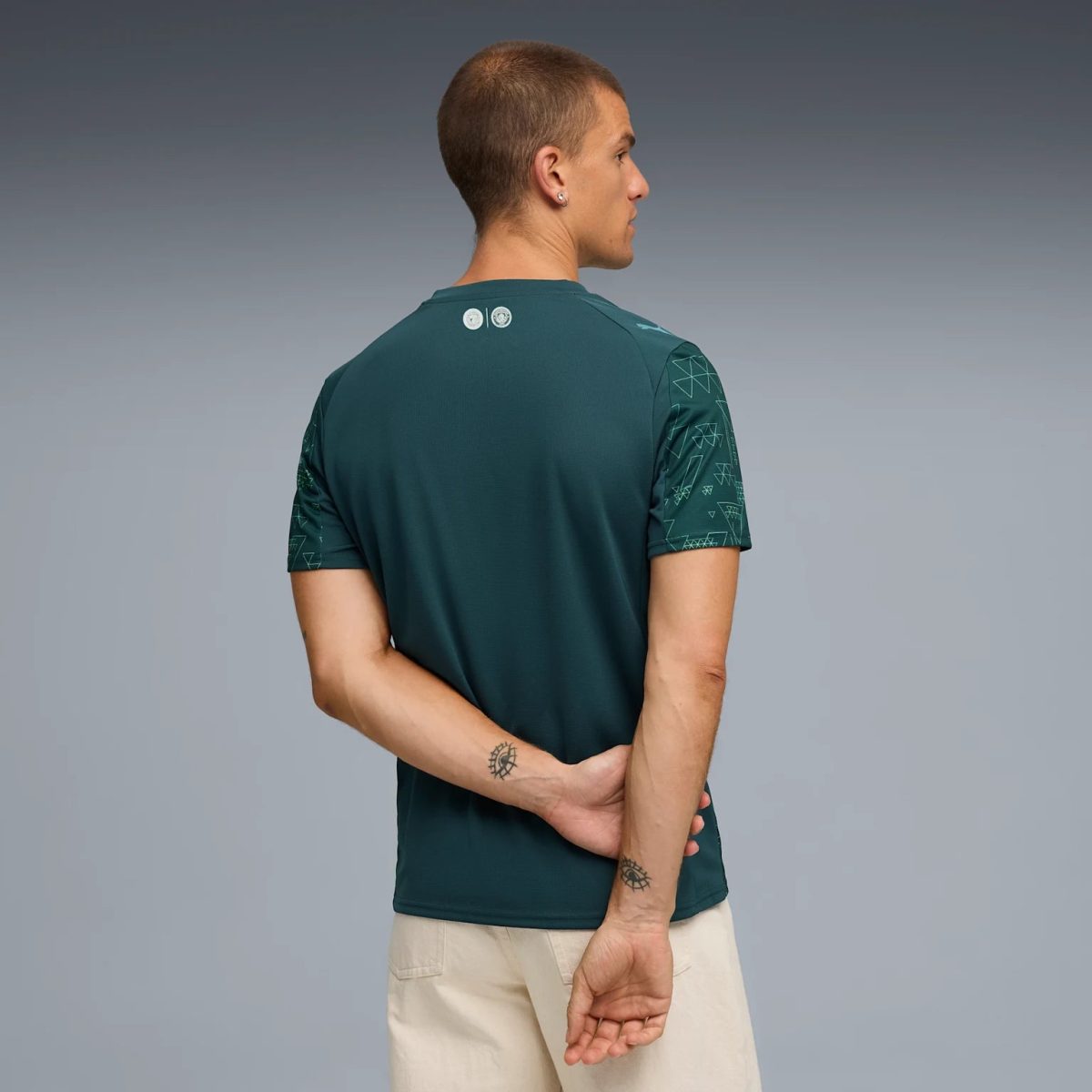 Back view of a man wearing a teal short-sleeve shirt with geometric sleeve patterns, hands loosely clasped, beige shorts, against a gray gradient background.