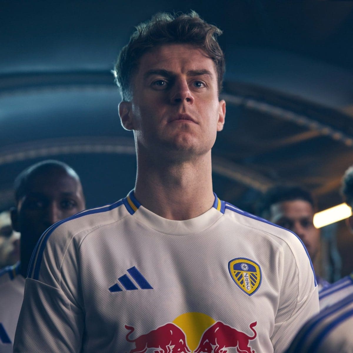 Male footballer in a Leeds United kit stands in a tunnel, looking forward with a determined expression.